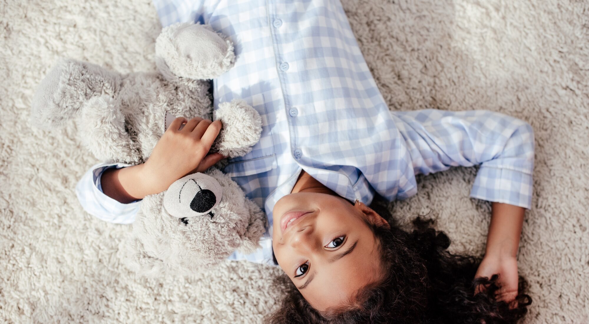 top view of adorable african american child in pajamas lying with teddy bear on carpet in room and